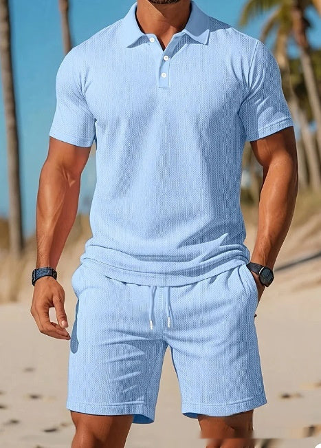 A man wearing a light blue ribbed knit polo shirt and matching drawstring shorts, standing on a sandy beach with palm trees and blue sky in the background.