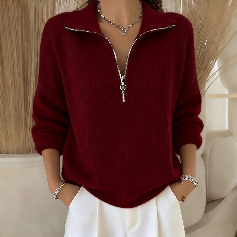 Person wearing a burgundy sweater with a zipper and white pants in a neutral setting