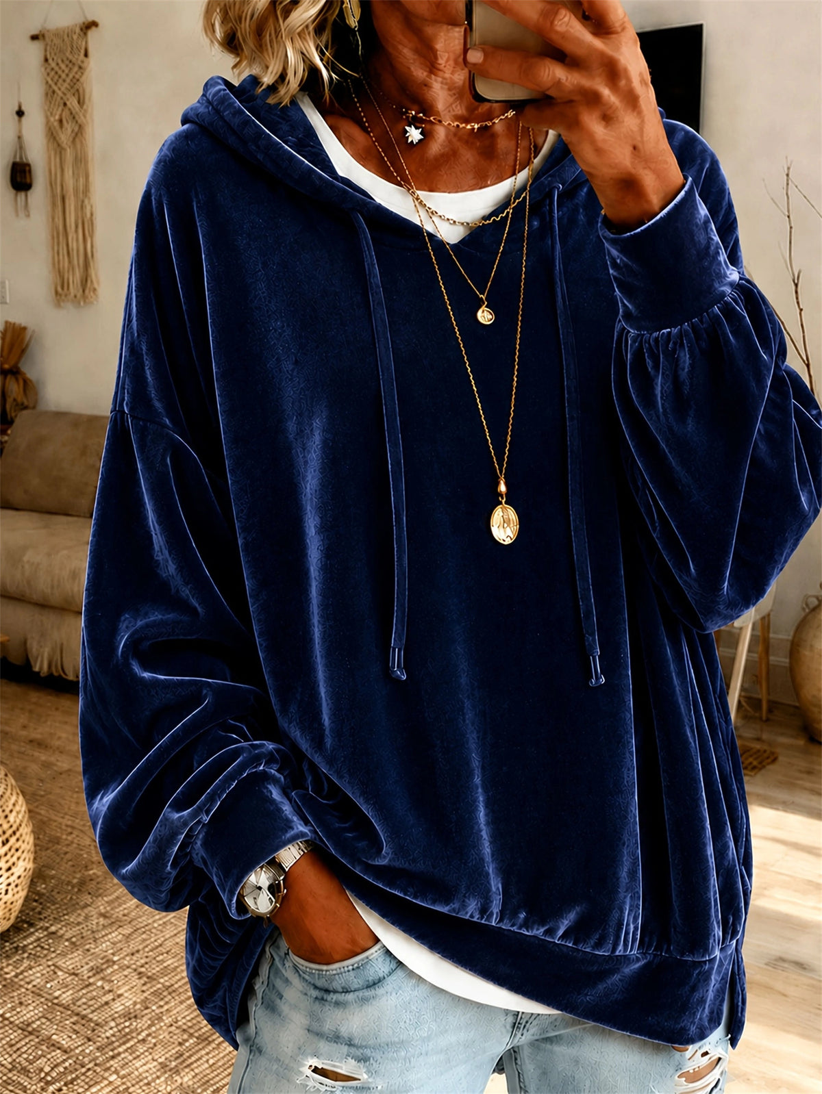 A person wearing an oversized navy blue velvet hoodie with drawstrings, layered over a white undershirt and light blue distressed jeans, photographed in a home interior setting.