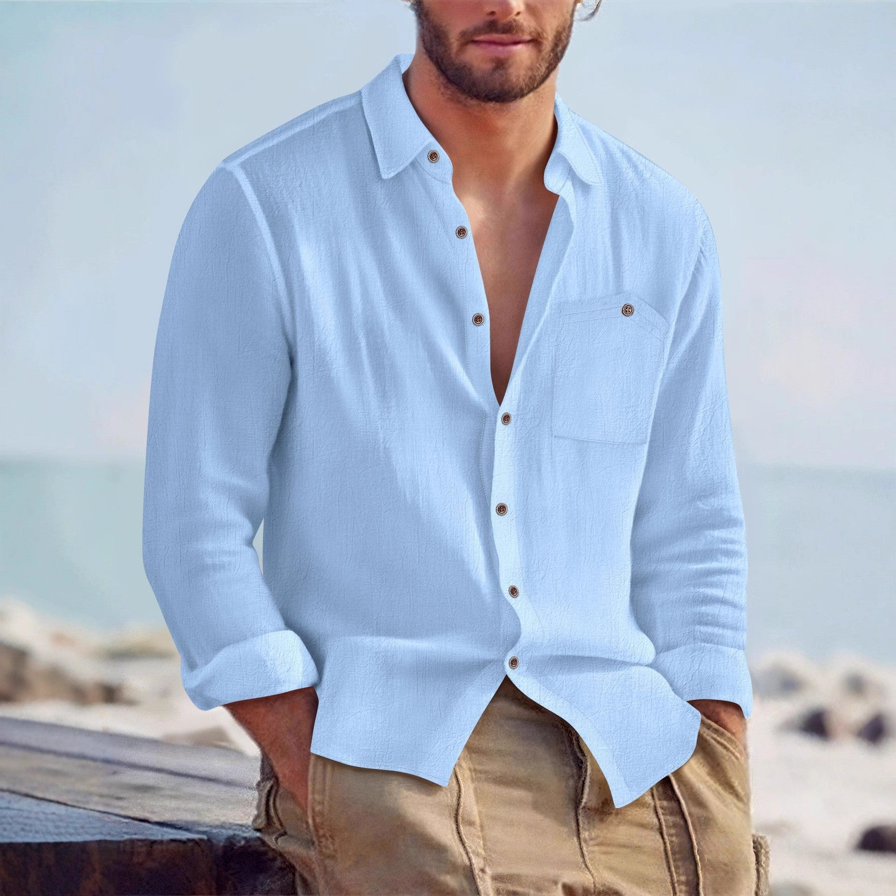 Man wearing a light blue shirt with rolled-up sleeves and beige pants by the beach.