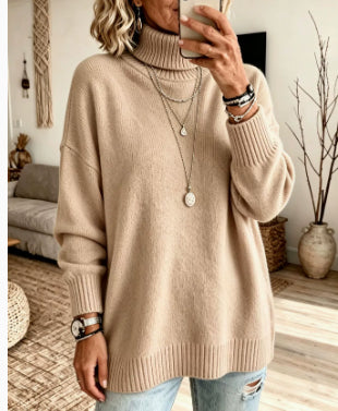 A woman wearing an oversized beige ribbed turtleneck sweater, paired with light blue distressed jeans and layered silver necklaces, taking a mirror selfie in a cozy living room setting.
