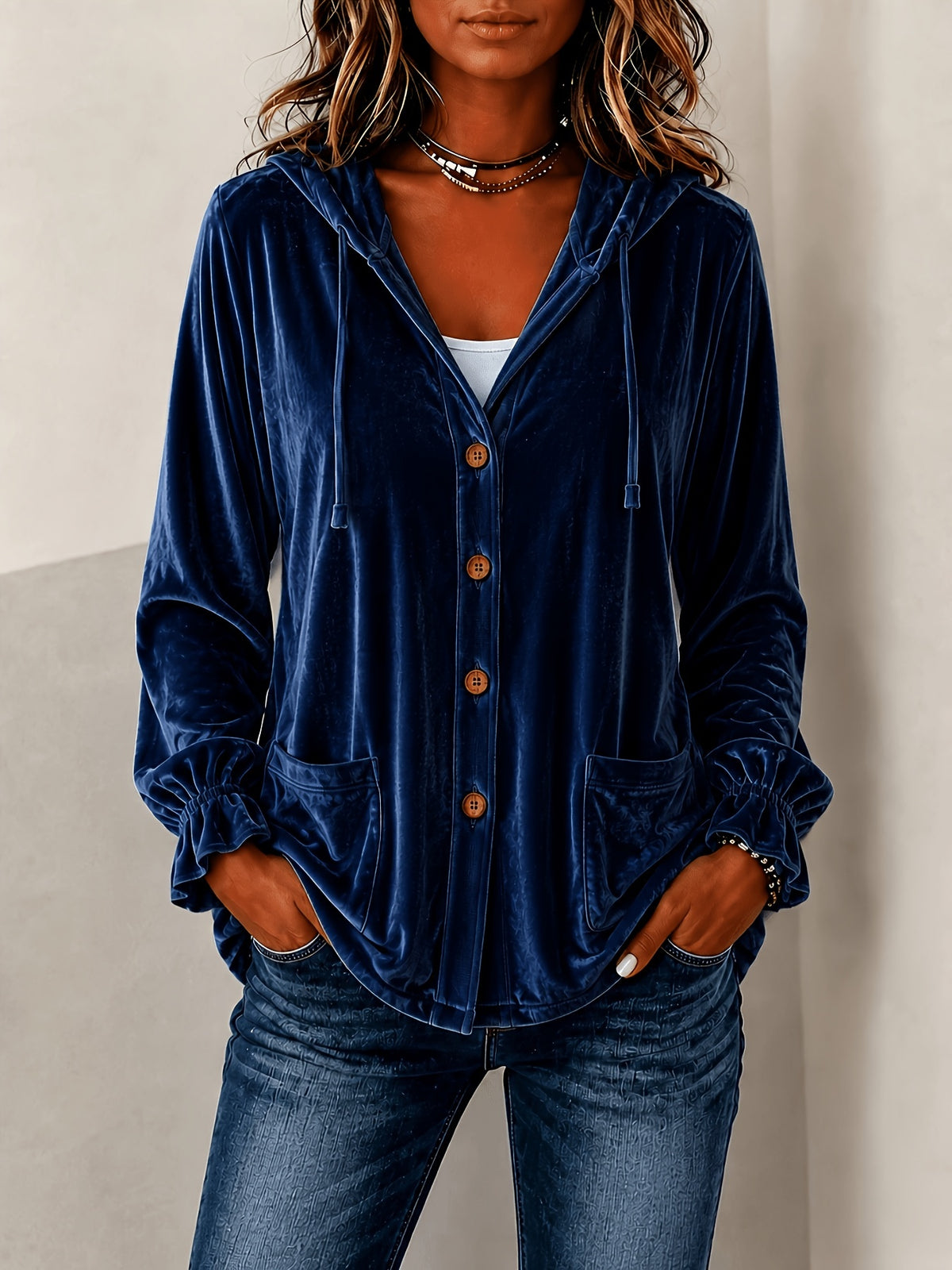 A woman wearing a navy blue velvet button-up hooded cardigan with front pockets, paired with blue jeans and layered necklaces, standing against a neutral beige wall.