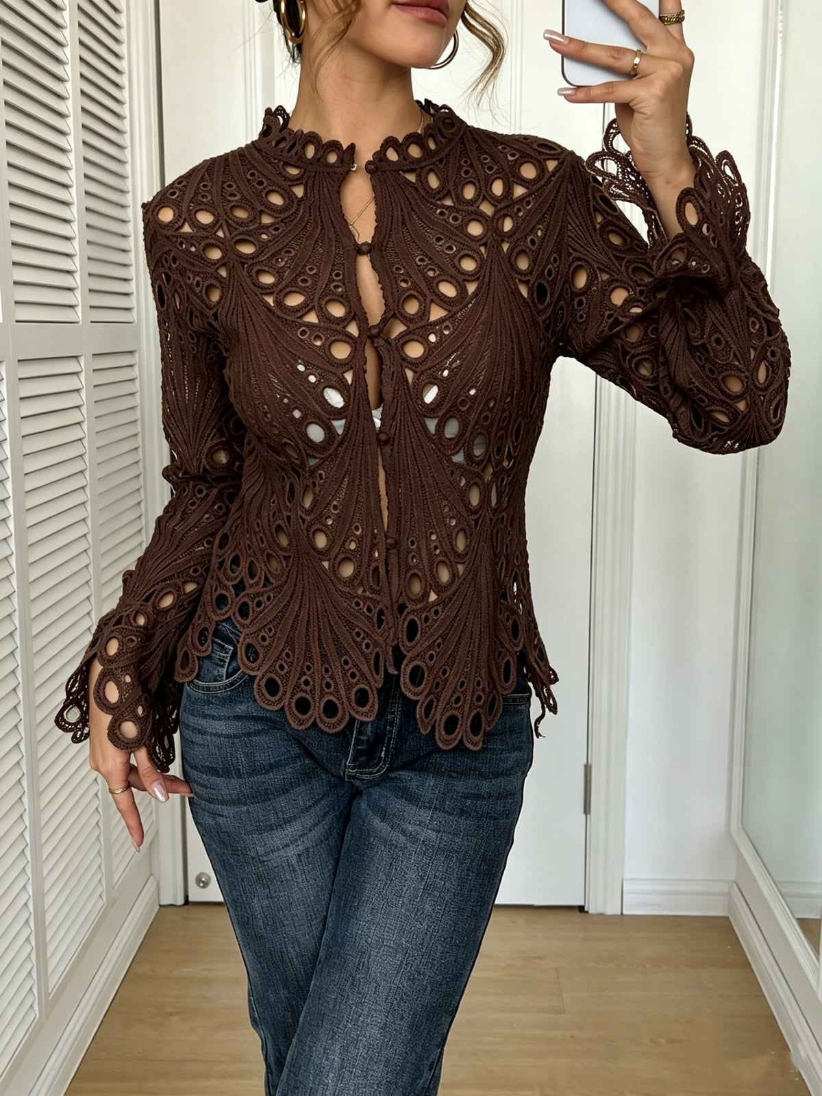 A woman wearing a brown crochet lace long-sleeve top with a keyhole cutout and blue jeans, taking a mirror selfie in an indoor room with white doors.
