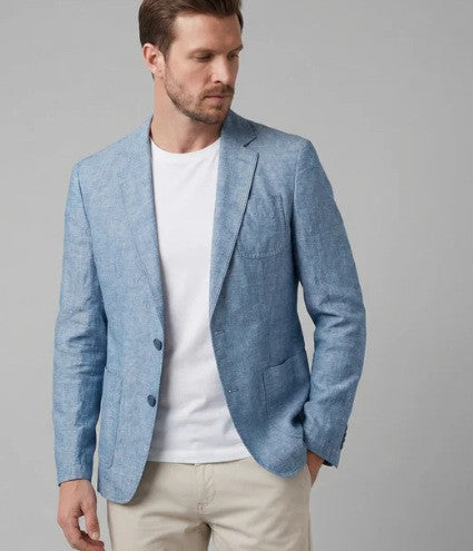 A man wearing a light blue textured blazer over a white t-shirt and beige trousers, photographed against a solid gray studio background.