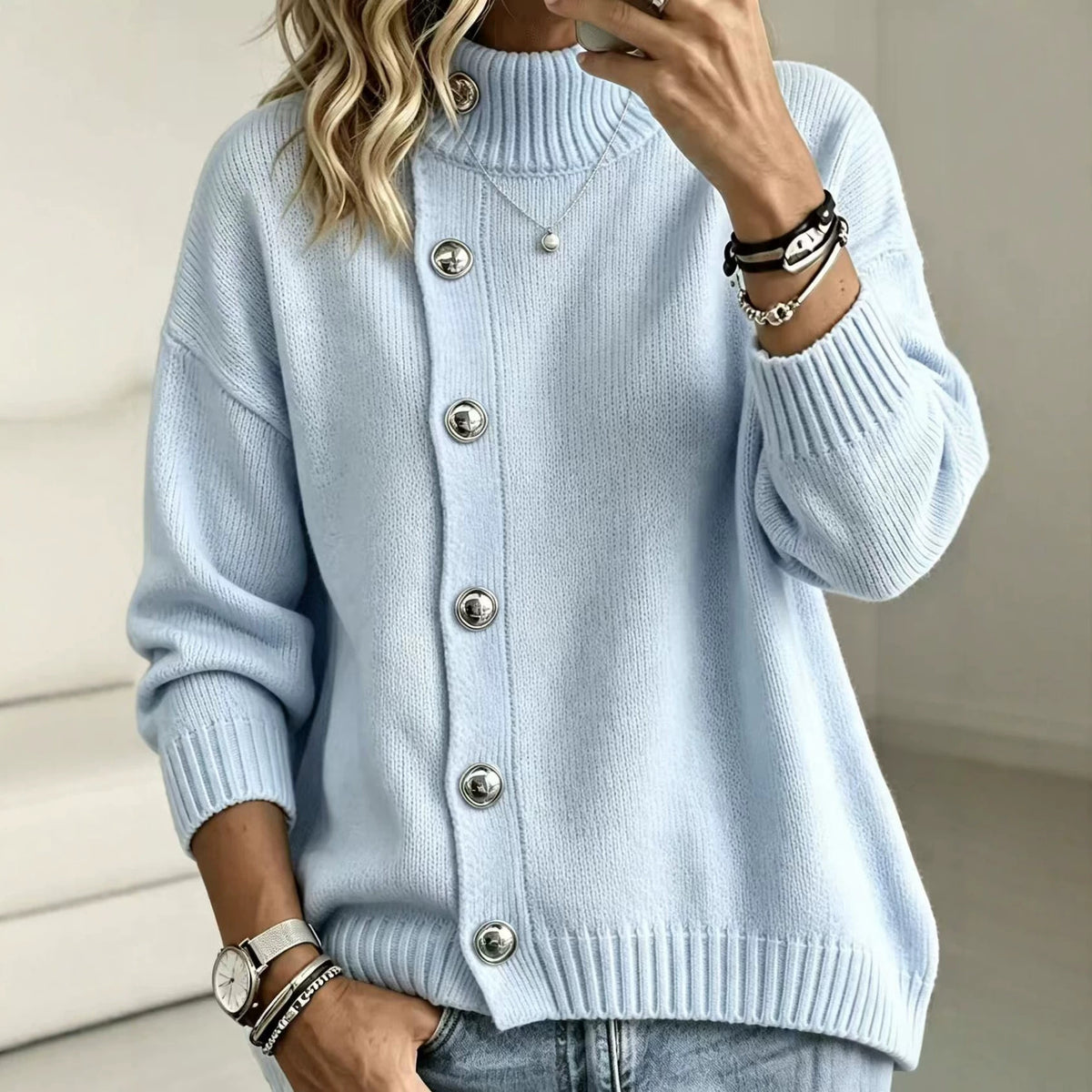 A woman wearing a light blue ribbed knit cardigan sweater with large silver buttons, paired with light-wash jeans. She is shown in a mirror selfie style, wearing bracelets and a necklace.