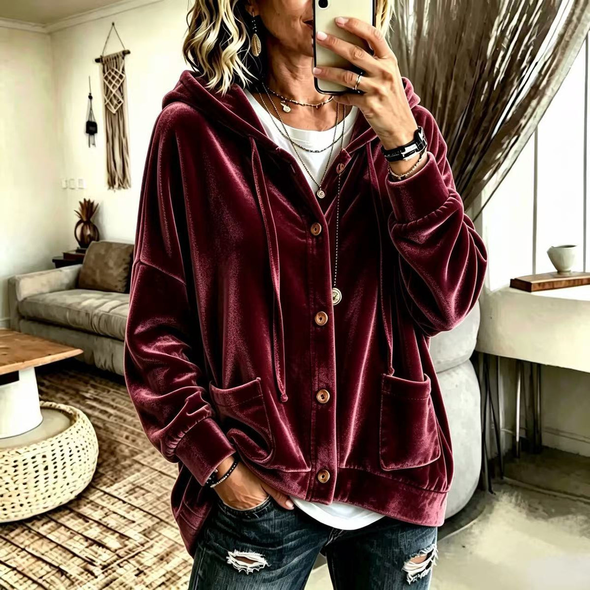 A woman takes a mirror selfie wearing a burgundy velvet zip-up hoodie jacket with a hood, over a white t-shirt and distressed dark jeans in a cozy living room setting.