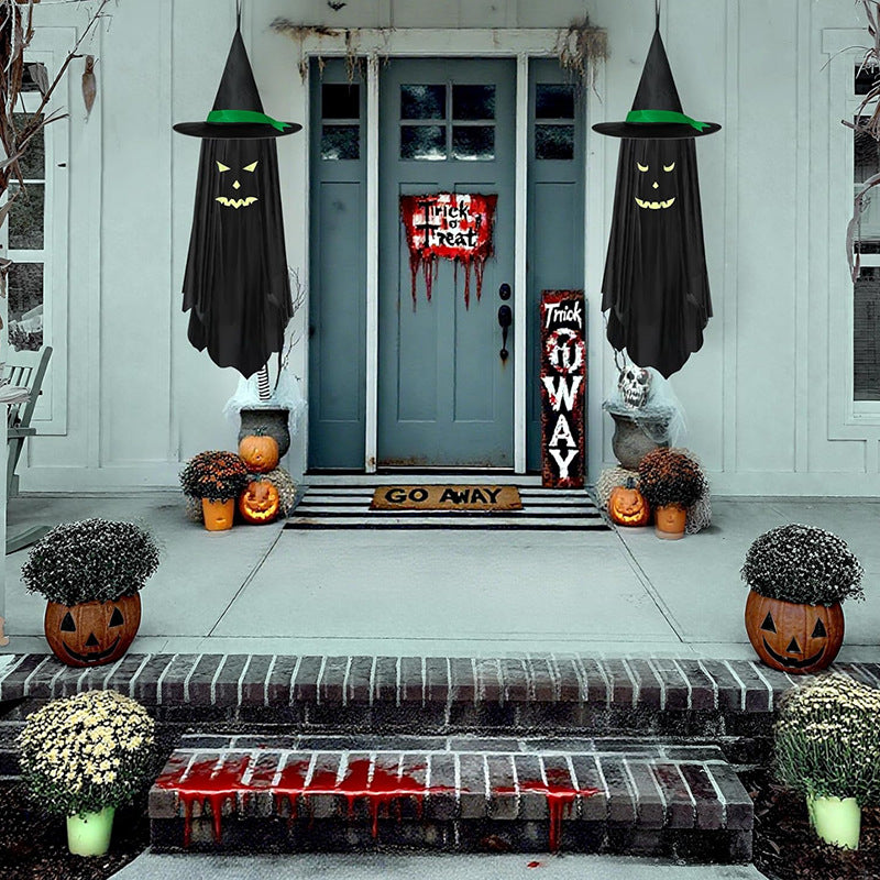 A front porch decorated for Halloween with two black witch cloaks hanging on either side of the door, a vertical 'Trick or Treat' sign, jack-o'-lanterns, potted flowers, and fake blood on the steps. The door has a 'Happy Halloween' sign with dripping red paint.