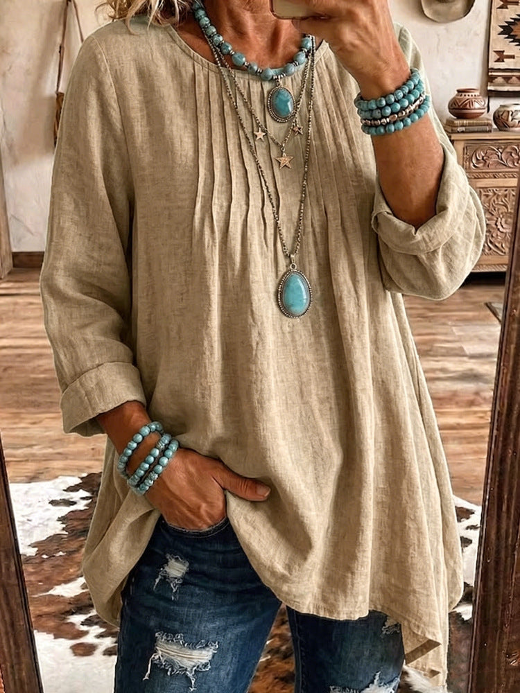 A woman takes a mirror selfie wearing a beige linen-blend tunic top with vertical pleats, paired with distressed blue jeans and layered turquoise jewelry.