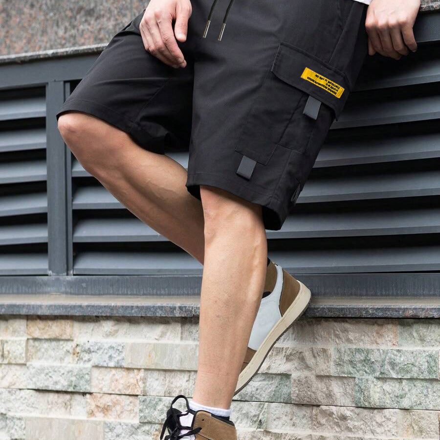 Person wearing black cargo shorts with a visible brand label, standing against a stone wall.