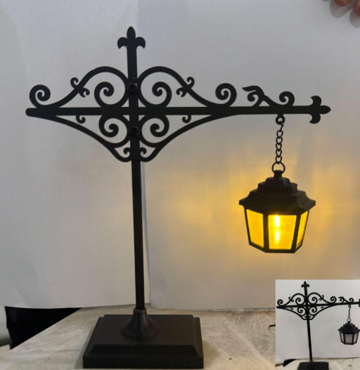 Black wrought iron decorative table lamp with a hanging lantern, shown lit against a plain light background. A small inset at bottom right shows the unlit product view.