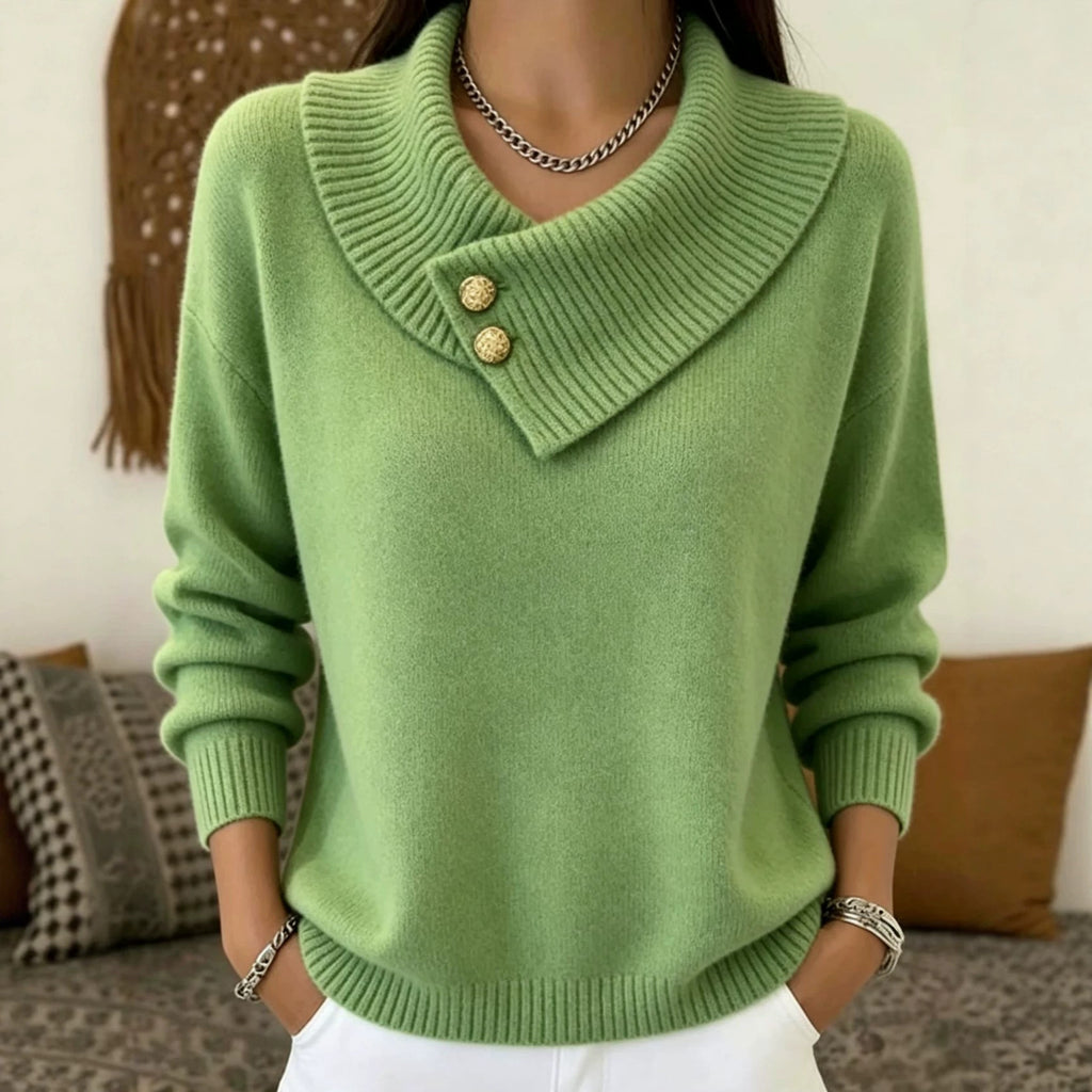 A woman wearing a light green knit sweater with a wide, folded collar and two decorative gold buttons, paired with white pants and jewelry, in a cozy indoor setting.