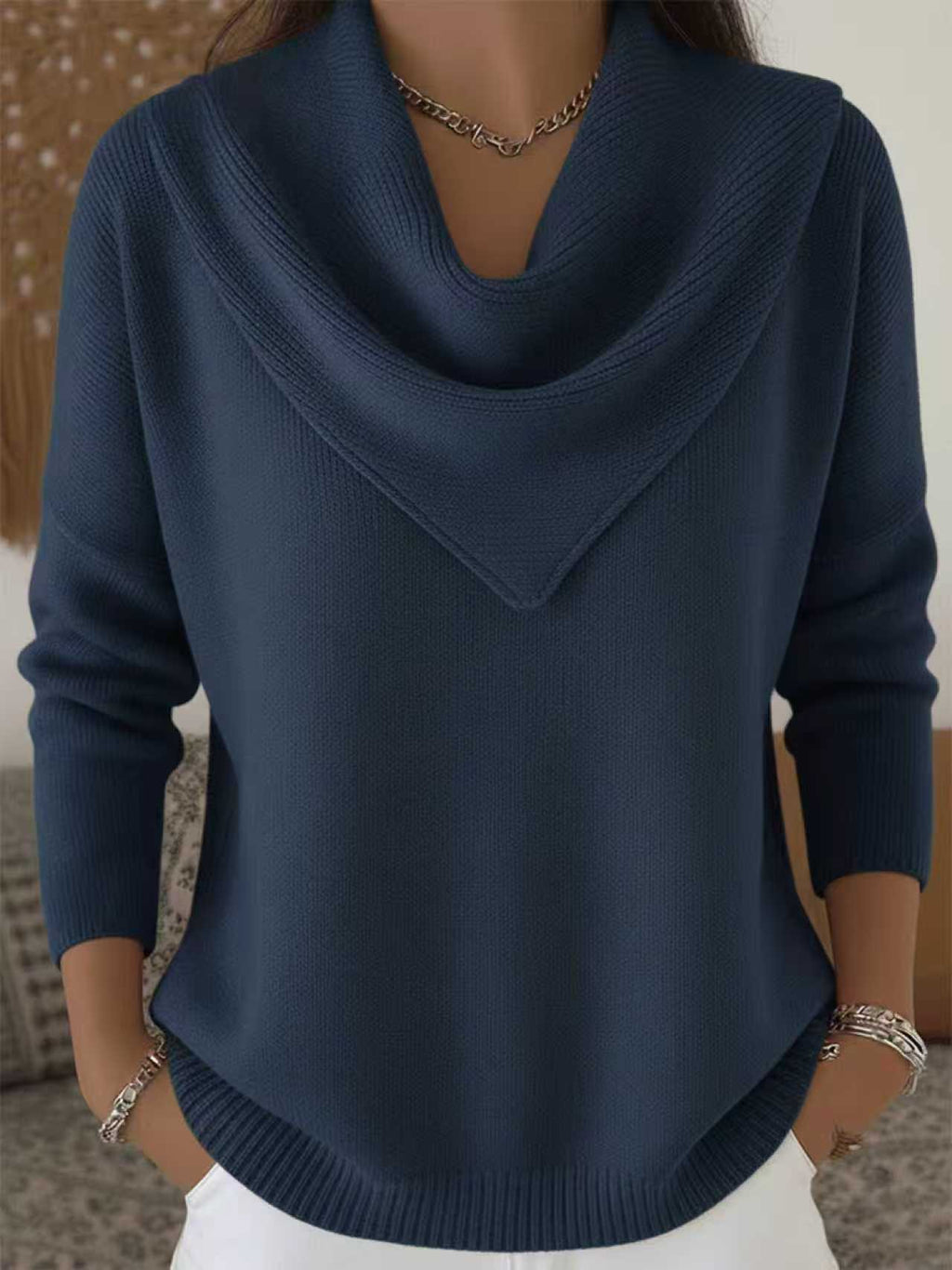 A close-up of a woman wearing a dark blue cowl-neck knit sweater with long sleeves, paired with white pants and silver jewelry.