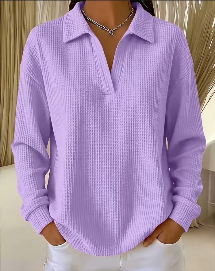 A woman wearing a lavender purple waffle-knit long-sleeve polo shirt with white pants, standing indoors with her hands in her pockets.