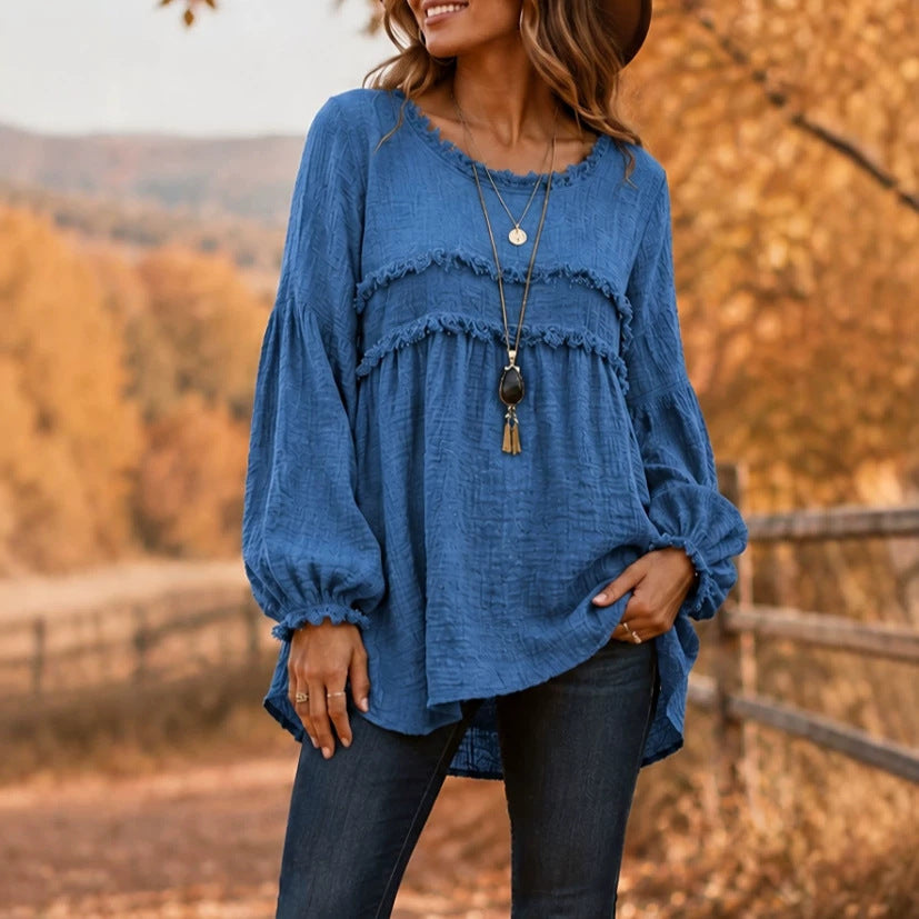 A woman wearing a blue long-sleeve blouse with a frayed ruffle trim and gathered waist, paired with dark jeans, standing outdoors in front of a blurred autumn landscape.