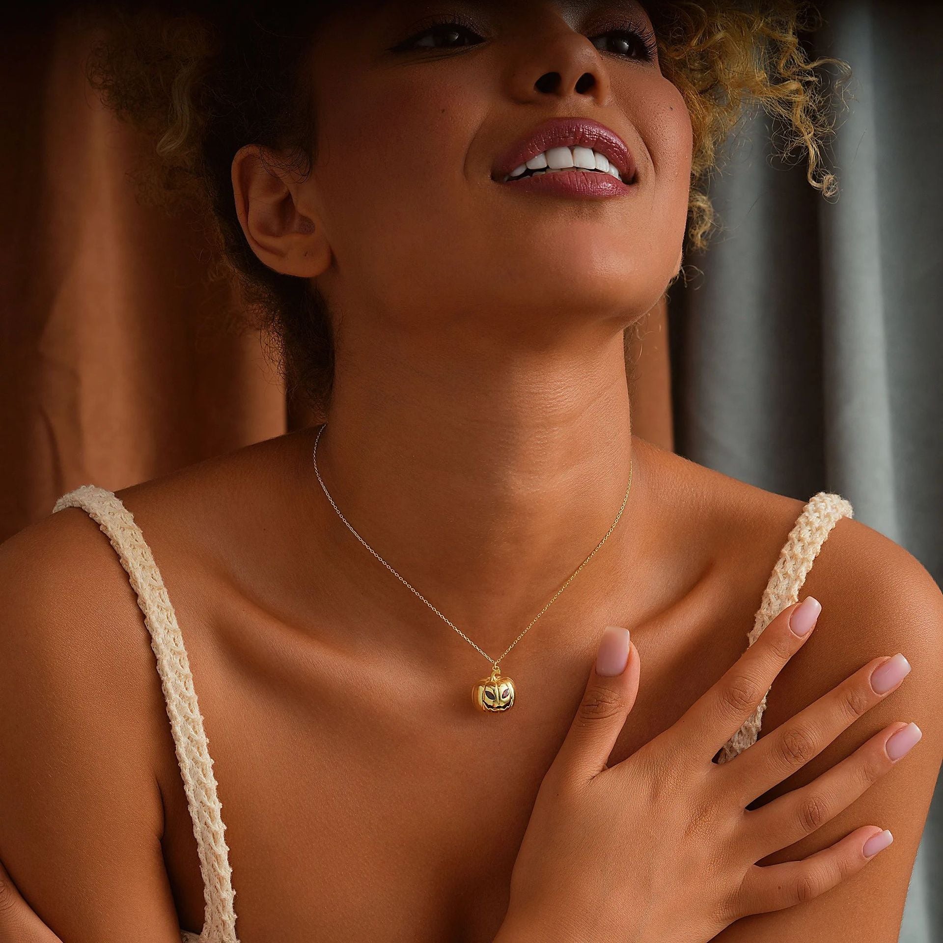 A woman wearing a delicate gold chain necklace with a small gold heart pendant, styled with a cream-colored crochet camisole against a warm brown and gray curtain background.