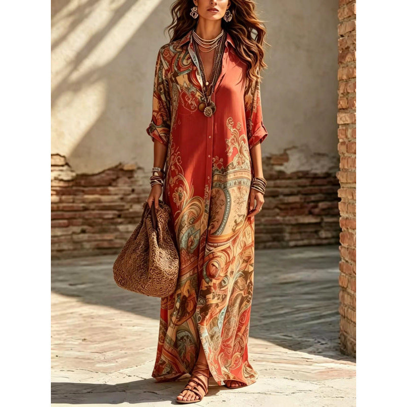 A woman models a bohemian-style maxi shirt dress with a colorful paisley print in rust orange, beige, and teal. She is standing in an outdoor rustic setting with a brick column and textured wall, holding a woven straw tote bag.