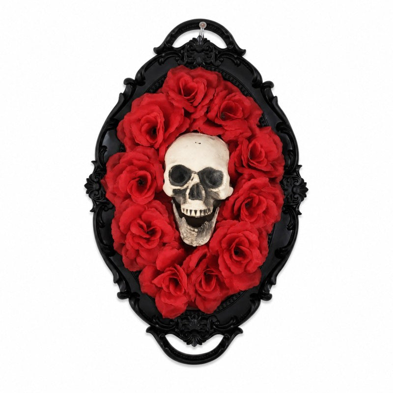 A gothic-style oval wall hanging featuring a realistic skull centered within a circular arrangement of vibrant red roses, all set inside an ornate black frame with decorative scrollwork and hanging loops.