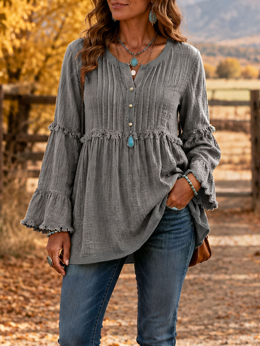A woman wearing a gray long-sleeve blouse with a buttoned V-neck, gathered waist, and ruffled cuffs, paired with blue jeans in an outdoor autumn setting.