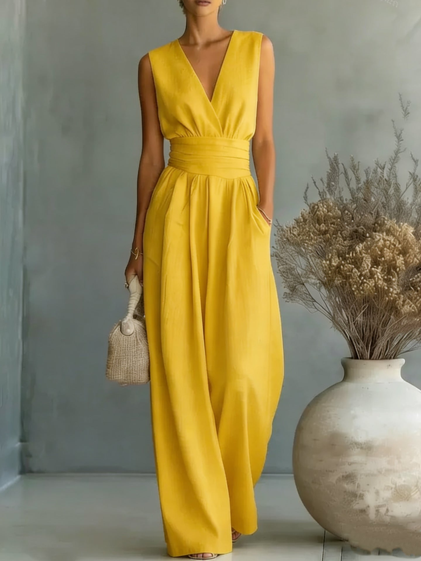 Woman wearing a yellow jumpsuit standing next to a vase with dried plants against a gray wall.