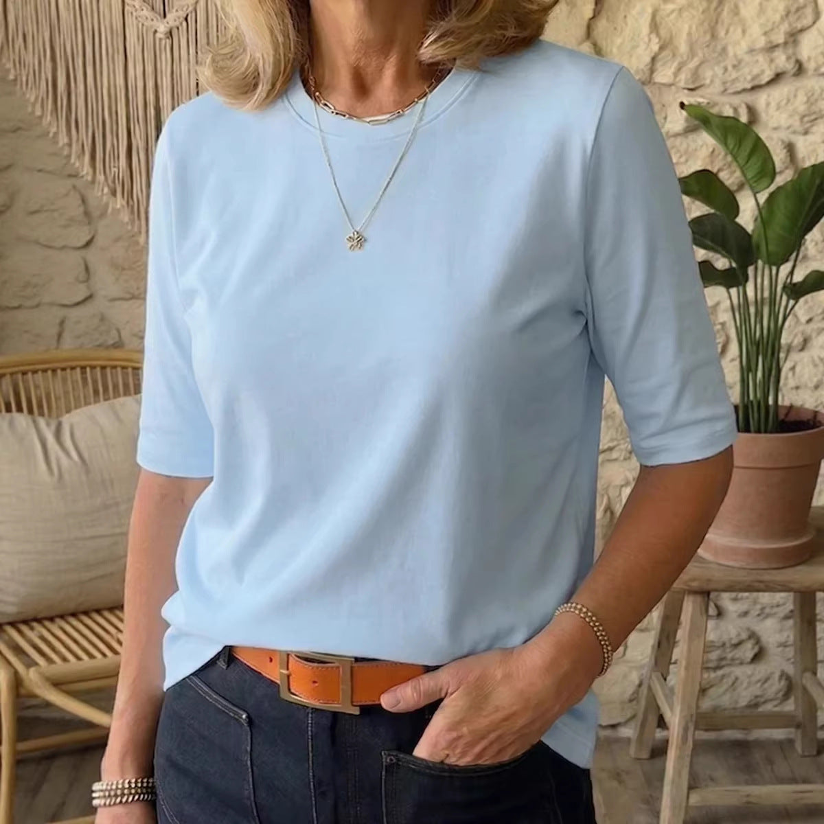 A woman wearing a light blue short-sleeve crewneck t-shirt, styled with dark blue jeans, a brown belt, and layered gold necklaces, standing in a rustic room with a stone wall and a potted plant.
