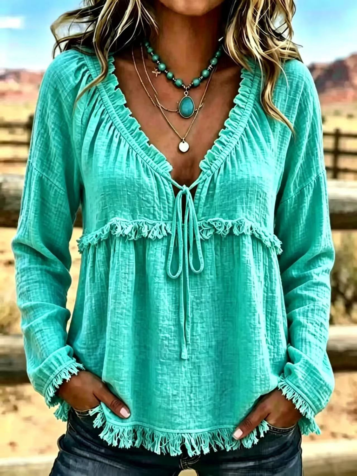 A woman wearing a long-sleeve turquoise green bohemian-style blouse with ruffles, a tie-front neckline, and fringe trim, paired with layered turquoise necklaces and dark jeans, set against a blurred outdoor desert landscape.