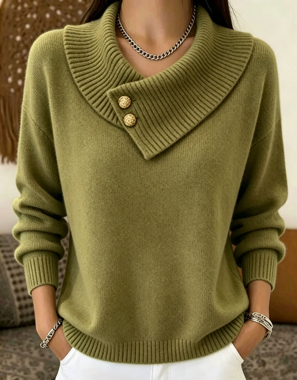 A woman wearing an olive green knit sweater with a wide folded collar and two decorative gold buttons, paired with white pants and silver jewelry.