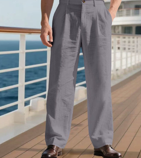 A person wearing light gray wide-leg trousers and dark brown leather dress shoes, standing on a wooden deck with a white railing and ocean view in the background.