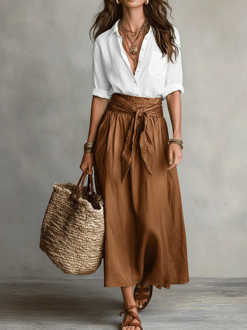Woman wearing a white blouse and brown skirt holding a woven bag against a neutral background