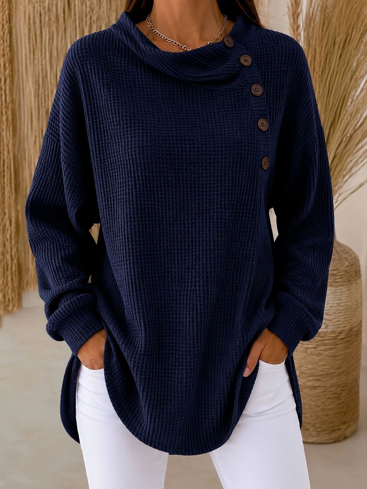 A woman wearing a navy blue waffle-knit long-sleeve top with an asymmetrical buttoned cowl neckline and white pants, standing indoors against a neutral background with dried grass decor.