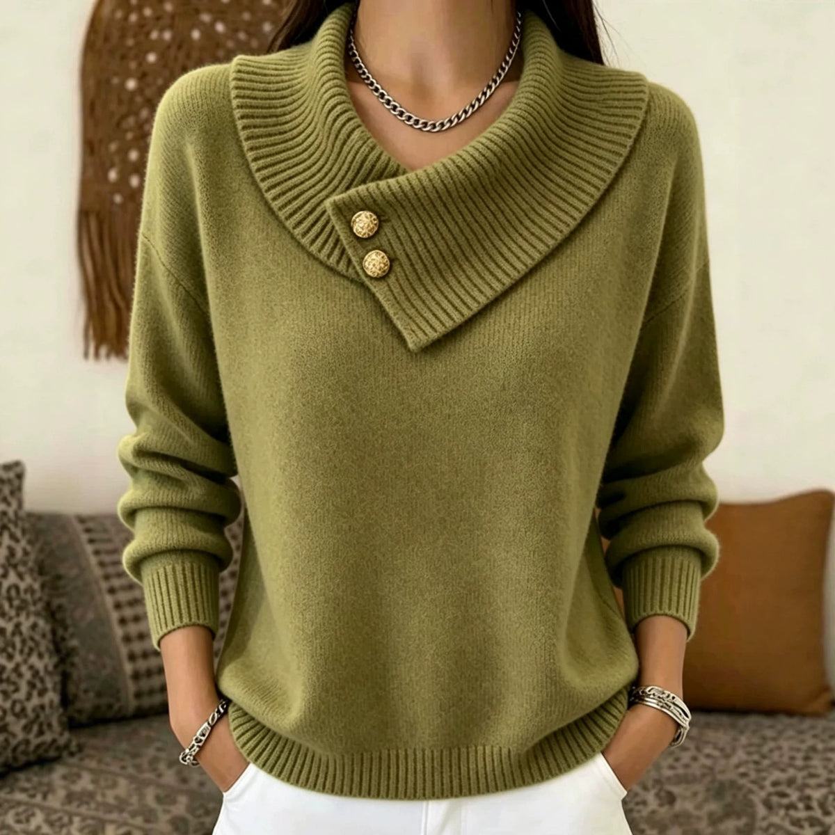 A woman wearing a ribbed knit sweater with a wide asymmetrical collar and two decorative gold buttons, paired with white pants and jewelry, in a softly lit indoor setting.