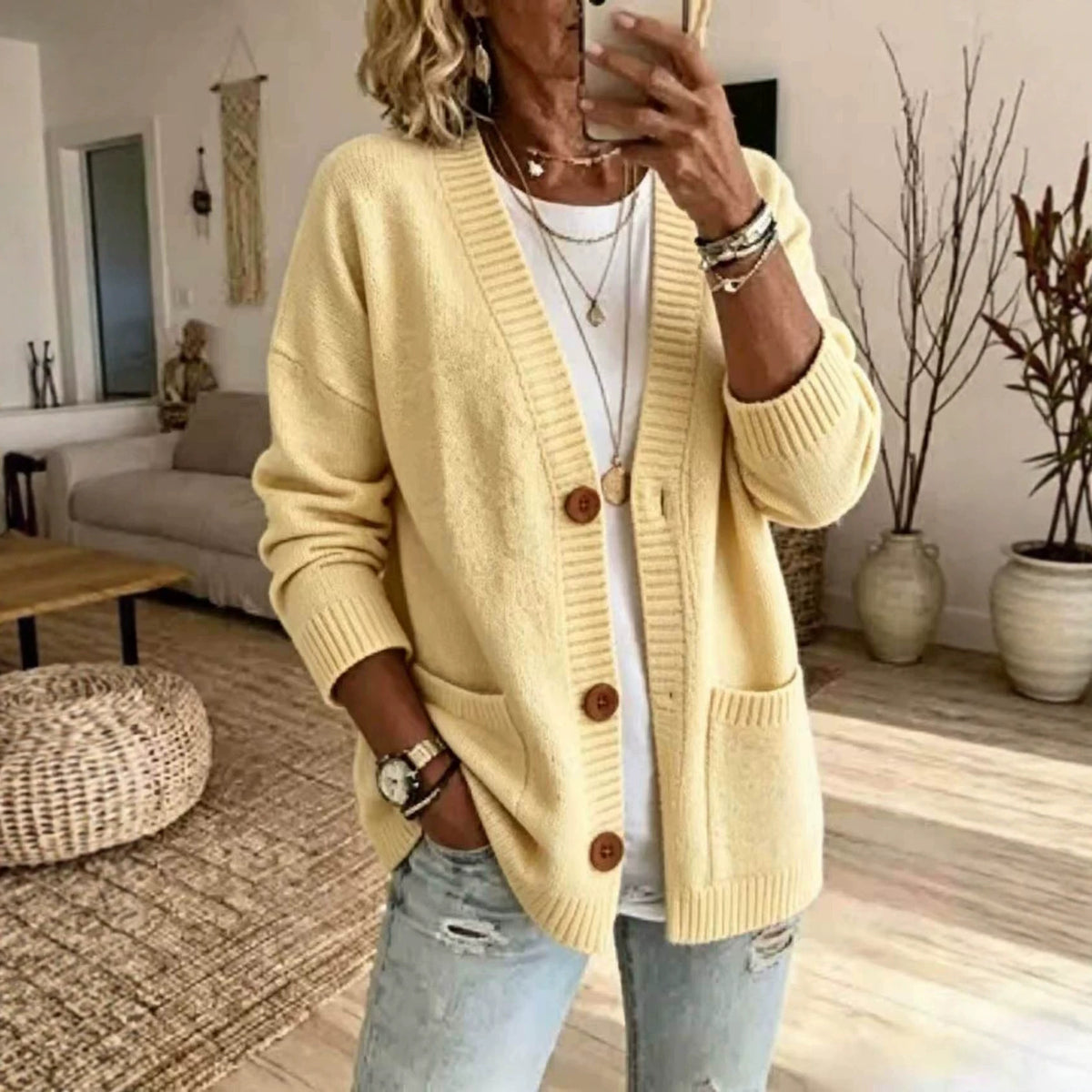 A woman takes a mirror selfie in a cozy living room, wearing a pale yellow knit cardigan with brown buttons over a white t-shirt and light-wash distressed jeans.