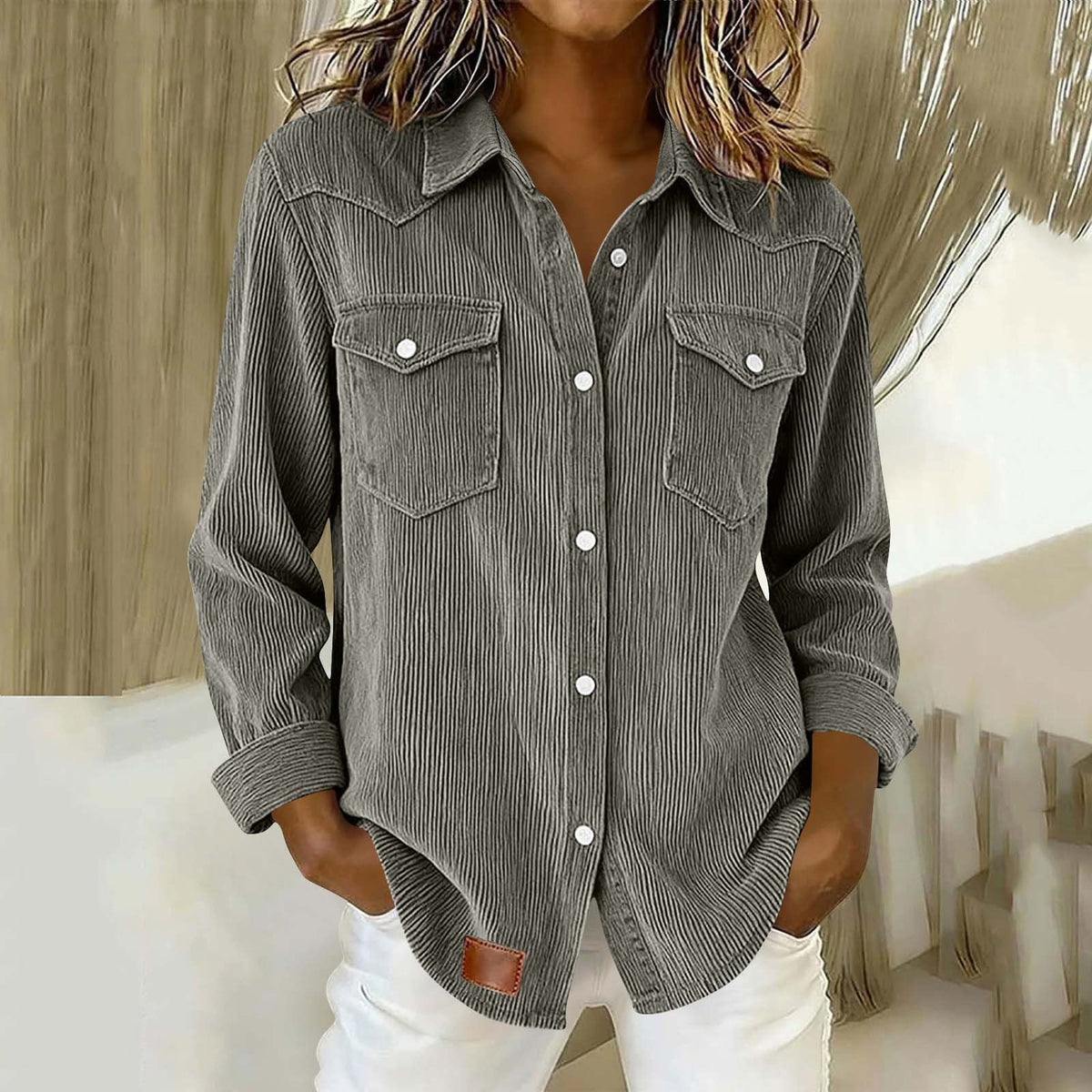 A woman wearing a dark gray corduroy button-up shirt with two chest pockets and white pants, standing in an indoor setting with a neutral background.