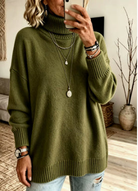 A woman wearing an oversized olive green ribbed turtleneck sweater, paired with light blue distressed jeans and layered silver necklaces, taking a mirror selfie in a living room setting.