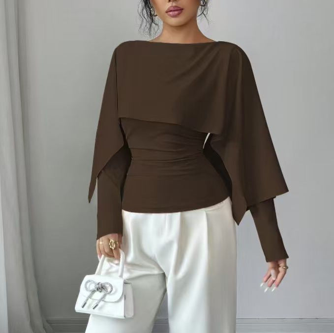 A woman wearing a dark brown long-sleeve top with a draped, cape-like layered overlay and gathered waist detail, paired with white pants and holding a small white handbag.