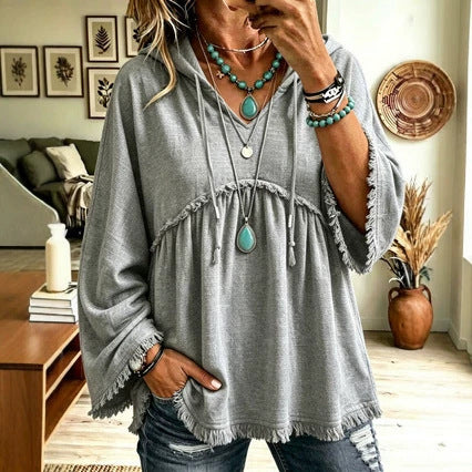 A woman wearing a loose, oversized gray bohemian-style tunic top with frayed edges and drawstring details, paired with distressed blue jeans and layered turquoise jewelry, in a cozy living room setting.