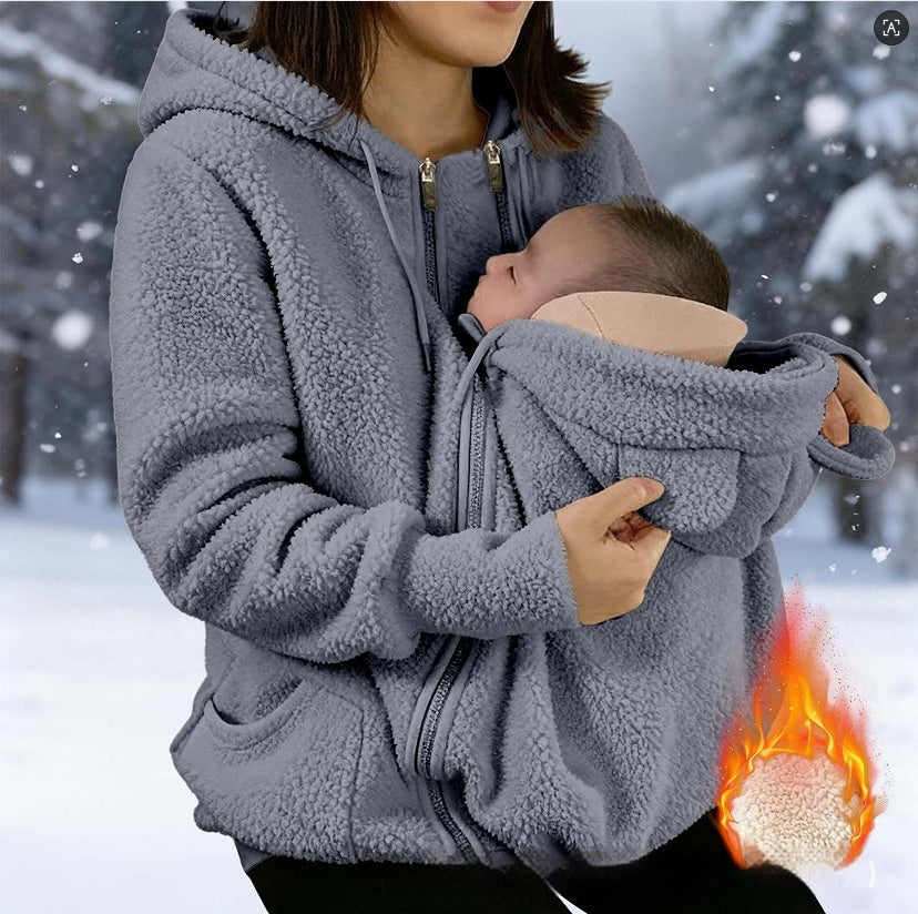 A woman in a gray sherpa zip-up hoodie carries a sleeping baby in a built-in hooded pocket, with a snowy outdoor background and a flame icon indicating warmth.