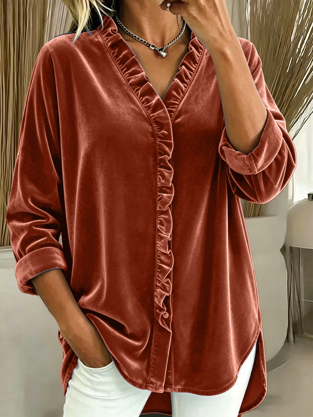A woman wearing a rust-colored velvet button-up blouse with ruffle trim along the front and collar, paired with white pants.
