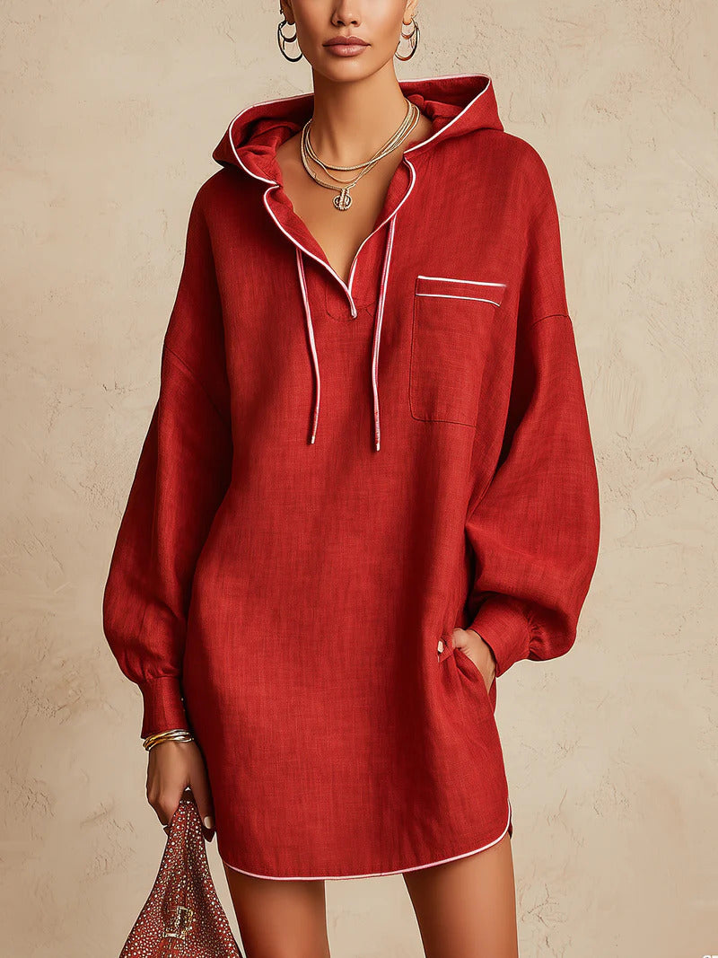 A woman models an oversized red hooded sweatshirt dress with white contrast piping, drawstrings, and a chest pocket, set against a textured beige background.