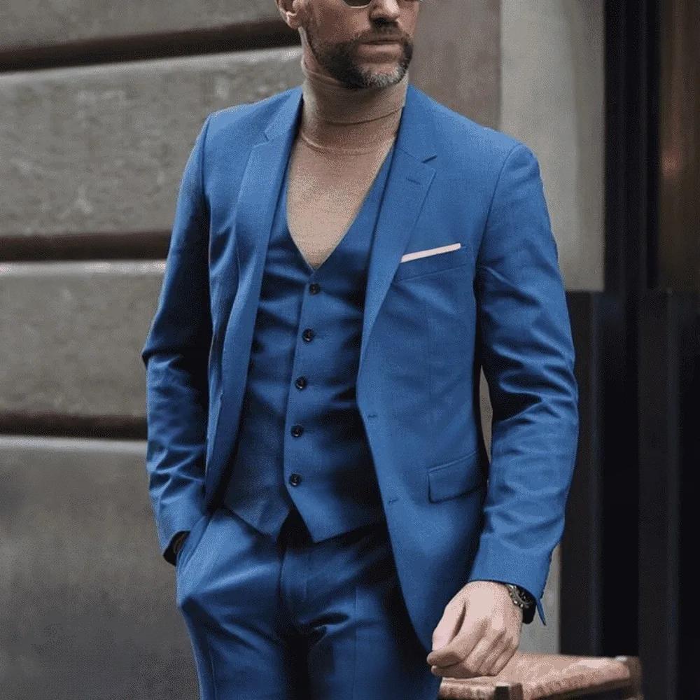 Man wearing a blue suit with a white shirt and black shoes, standing against a neutral background.