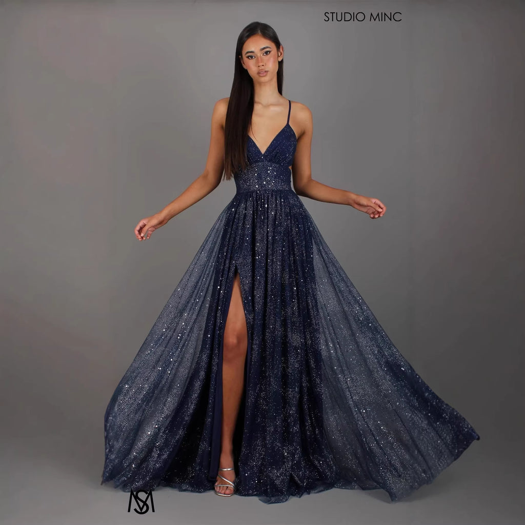 Woman wearing a sparkly navy blue evening gown with a slit on a gray background, branded 'Studio Minc'.