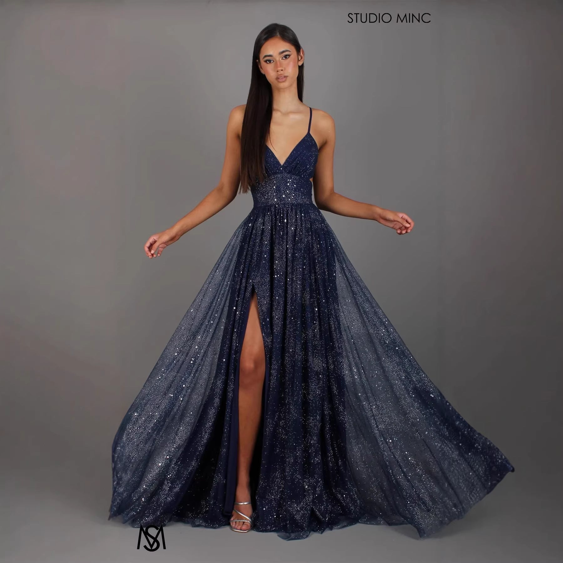 Woman wearing a sparkly navy blue evening gown with a slit on a gray background, branded 'Studio Minc'.
