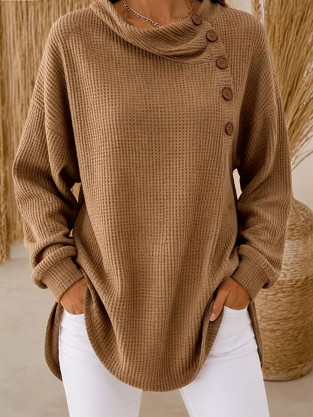 A woman wearing a brown waffle-knit cowl-neck sweater with a buttoned asymmetrical neckline, paired with white pants.