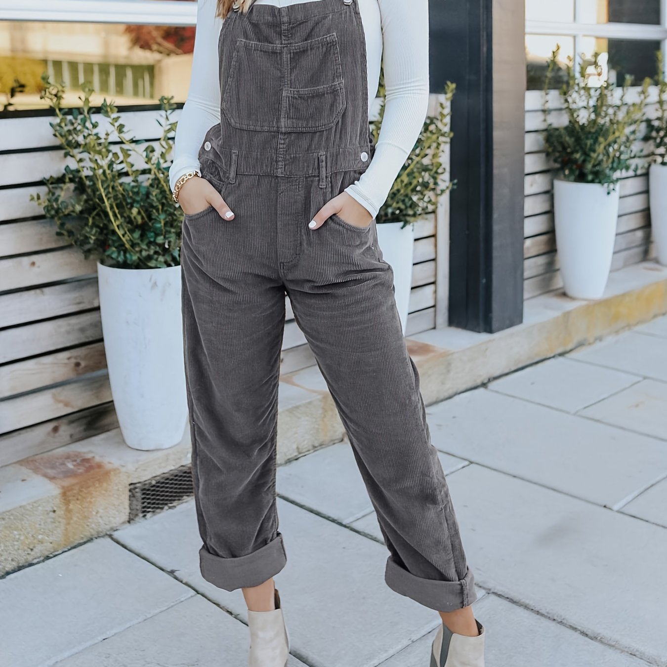 Person wearing gray overalls standing outdoors with plants and a building in the background