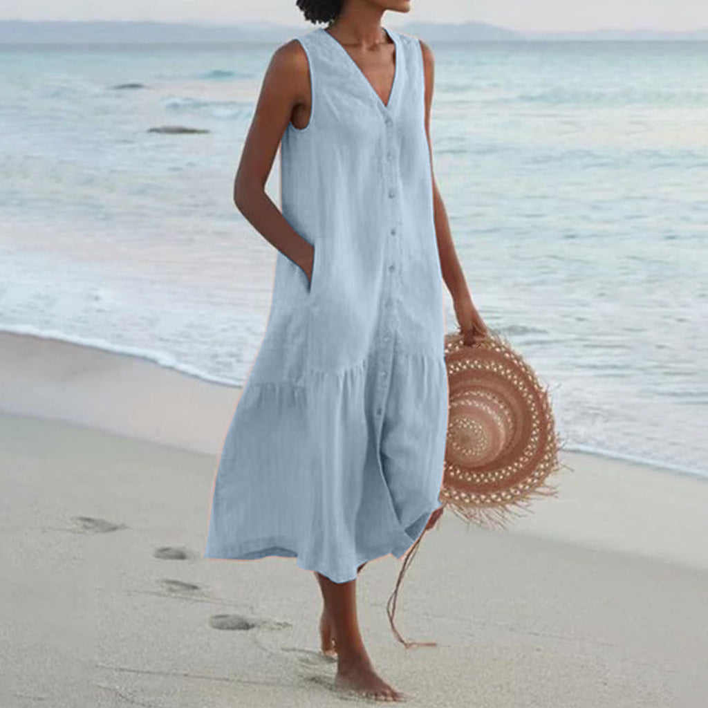 Woman in a light blue dress standing on a beach with a straw hat.