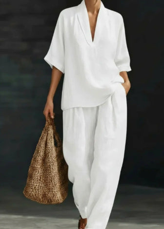 A model wearing a white two-piece linen outfit consisting of a short-sleeve V-neck top and matching wide-leg pants, holding a brown woven straw tote bag against a dark gray background.
