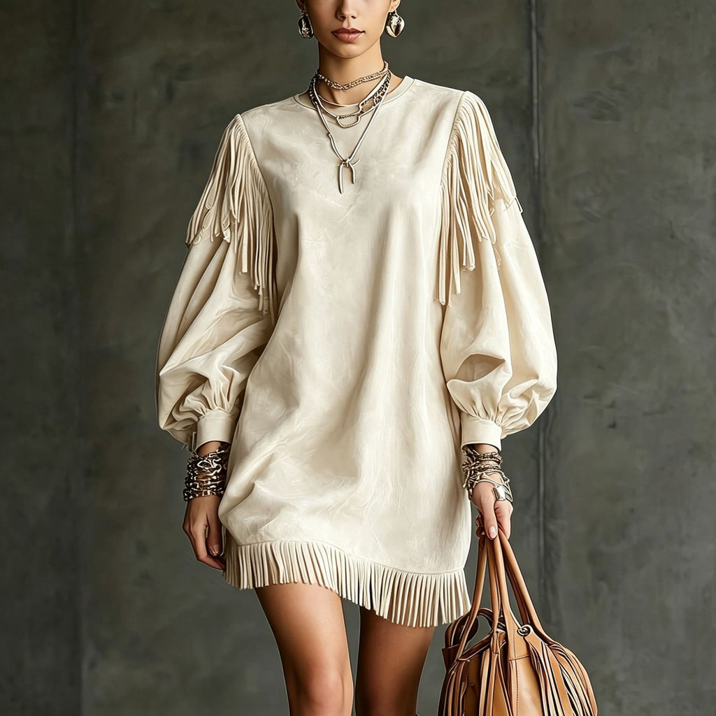 A model wears an oversized cream-colored faux-leather mini dress with long fringe detailing on the sleeves and hem, styled with layered necklaces, bracelets, and a brown leather handbag against a textured gray wall.