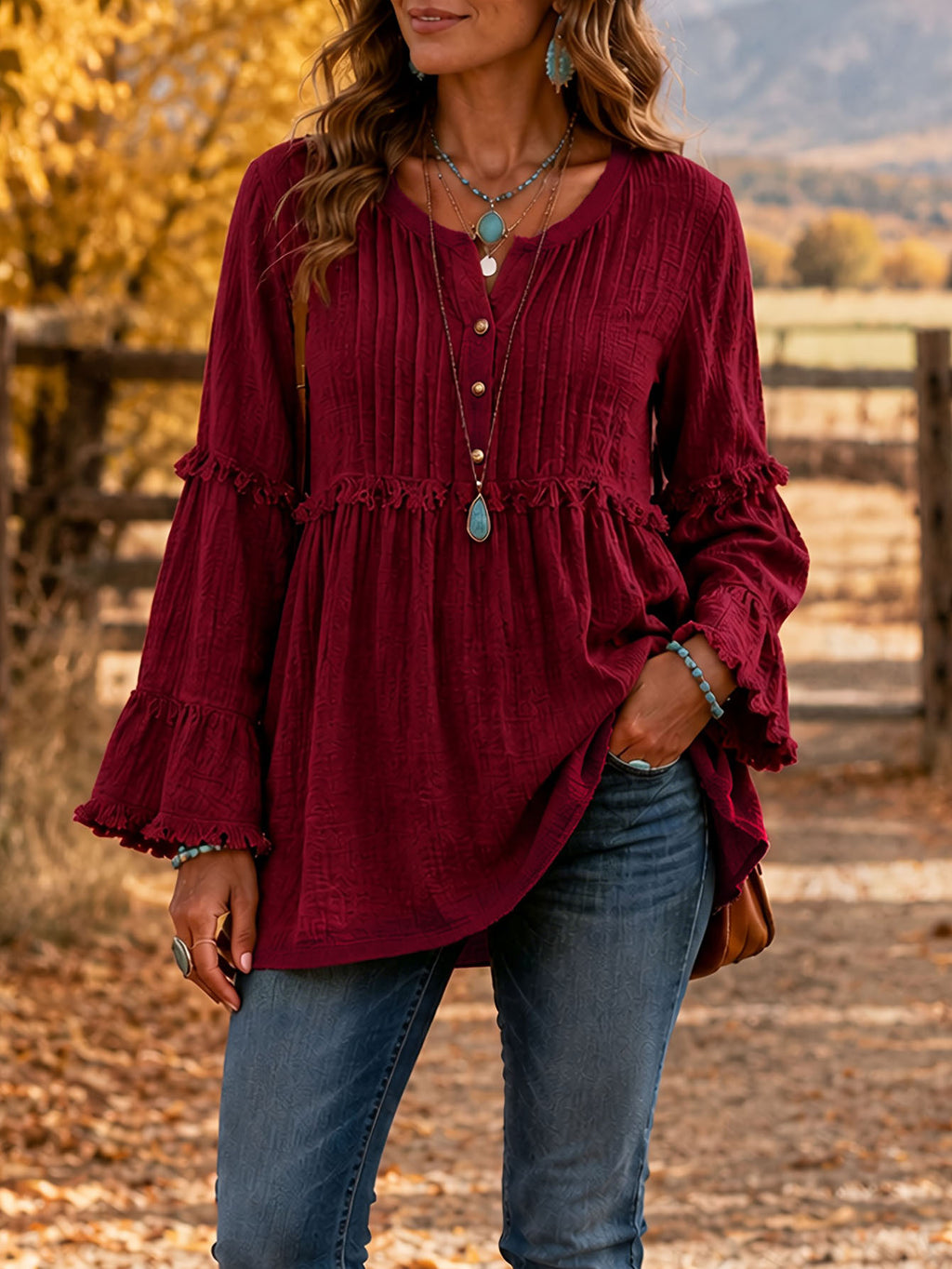 A woman wearing a long-sleeve burgundy top with a gathered peplum waist and ruffled cuffs, paired with blue jeans, standing outdoors in a fall setting.
