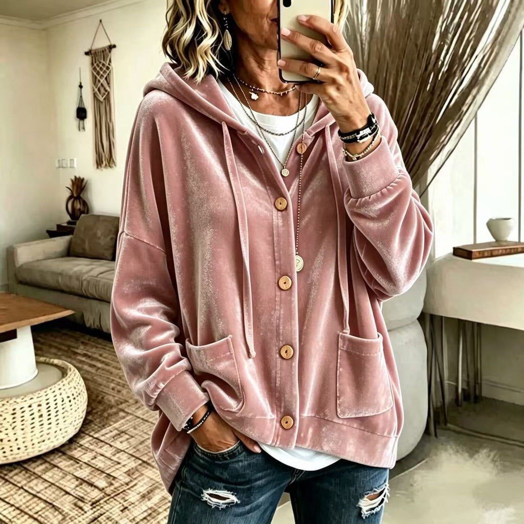 A woman takes a mirror selfie wearing a dusty pink velvet zip-up hoodie with drawstrings and front pockets, paired with distressed blue jeans in a cozy, modern living room setting.