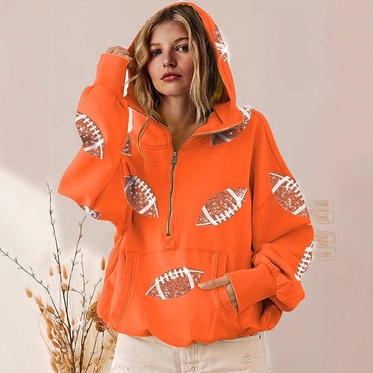 A woman wearing a bright orange half-zip hoodie with football print details, paired with off-white pants, standing against a light beige wall with dried plants in the foreground.