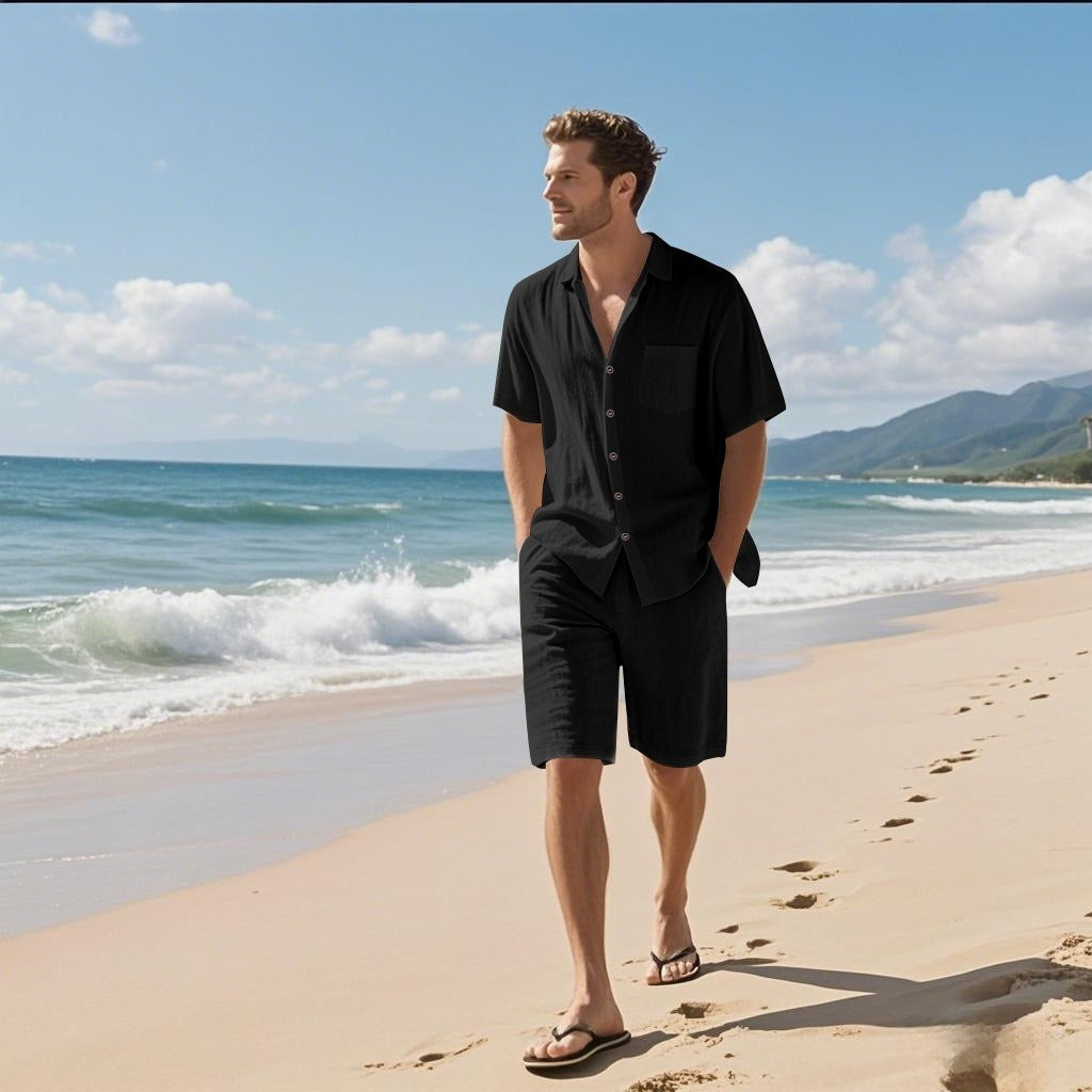 A man walks along a sandy beach wearing a black short-sleeve button-front shirt, black shorts, and black sandals, with ocean waves and a blue sky in the background.